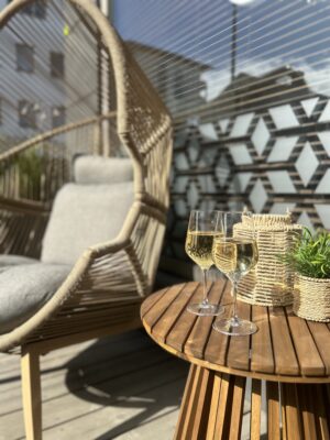 "Kaksi valkoviinilasia pyöreällä puupöydällä ulkoterassilla, taustalla rottinkinen riippukeinu ja moderni lasiseinä; kuva Ravintola Obelix Riihimäen terassitunnelmasta. Two glasses of white wine on a round wooden table on an outdoor terrace, with a hanging chair and modern glass wall in the background at Restaurant Obelix Riihimäki.