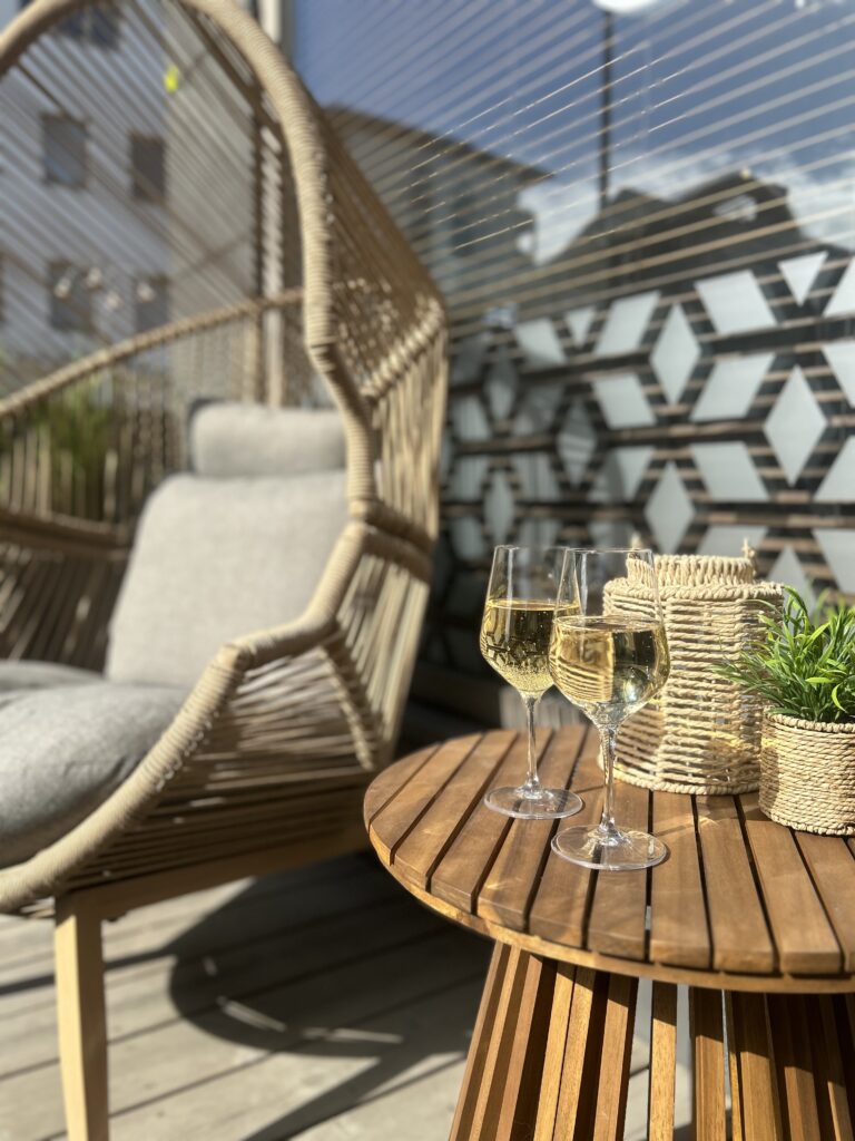 "Kaksi valkoviinilasia pyöreällä puupöydällä ulkoterassilla, taustalla rottinkinen riippukeinu ja moderni lasiseinä; kuva Ravintola Obelix Riihimäen terassitunnelmasta. Two glasses of white wine on a round wooden table on an outdoor terrace, with a hanging chair and modern glass wall in the background at Restaurant Obelix Riihimäki.