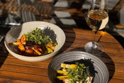 Kaksi kauniisti aseteltua annosta ja valkoviinilasi Ravintola Obelix Riihimäen terassilla, puupöydällä tarjoiltuna auringonvalossa, mukana paahdettuja kasviksia ja tuoreita versoja. Two beautifully plated dishes and a glass of white wine on the terrace of Restaurant Obelix in Riihimäki, served on a wooden table in warm sunlight with roasted vegetables and microgreens.