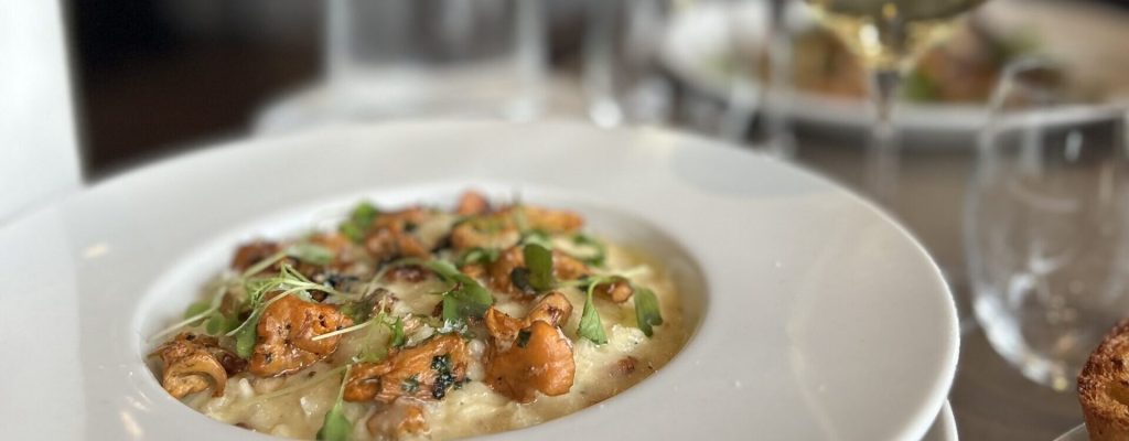 Valkoisessa kulhossa tarjoiltu kermainen risotto, jonka päällä paistettuja kantarelleja ja tuoreita yrttejä; taustalla punaviini‑ ja valkoviinilasi, vesikarahvi ja paahtoleipä erillisellä lautasella. Creamy risotto in a white bowl topped with sautéed mushrooms or seafood and fresh herbs; wine glasses, water carafe and toasted bread visible in the background.